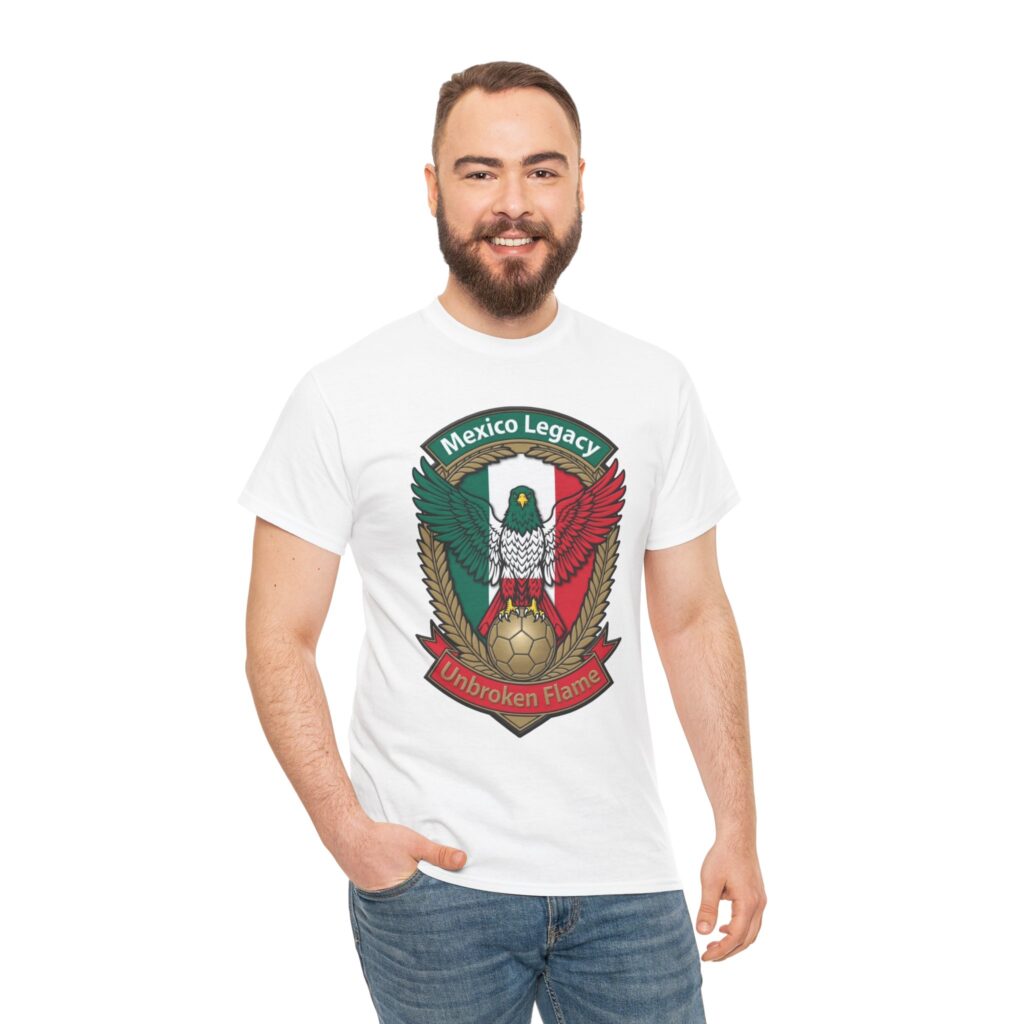 White t-shirt on a wooden hanger displaying a patriotic Mexican football emblem. A majestic green-headed eagle with spread wings stands atop a golden soccer ball, set against the Mexican flag (green-white-red vertical stripes). The design is framed by golden wheat/laurel branches, with a green top banner reading "Mexico Legacy" and a red bottom ribbon reading "Unbroken Flame". The overall style is bold, heraldic, and vibrant, celebrating Mexican national pride and soccer legacy.